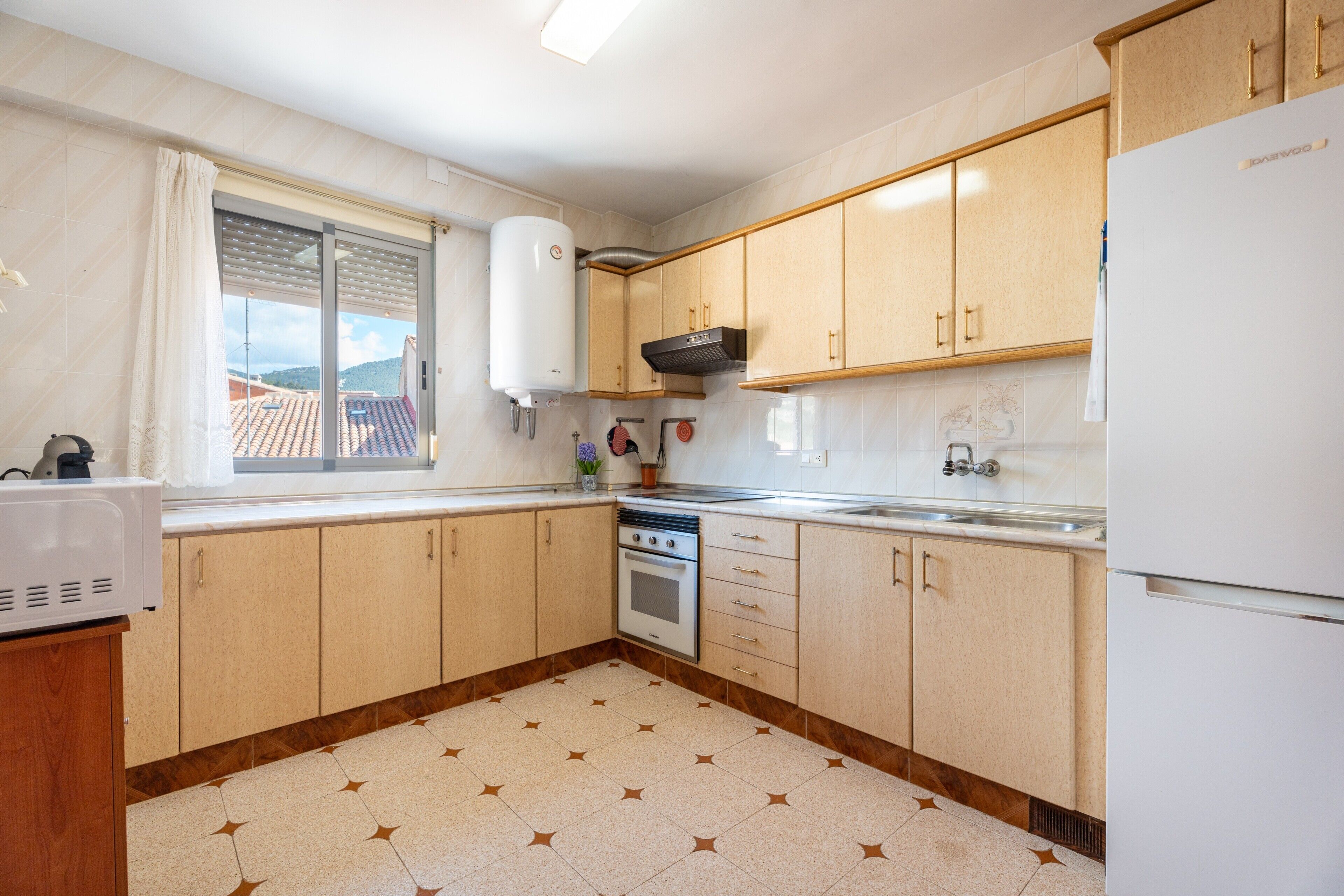 Une cuisine avec des meubles de rangement en bois, un four encastré, un réfrigérateur et une fenêtre donnant sur une maison.