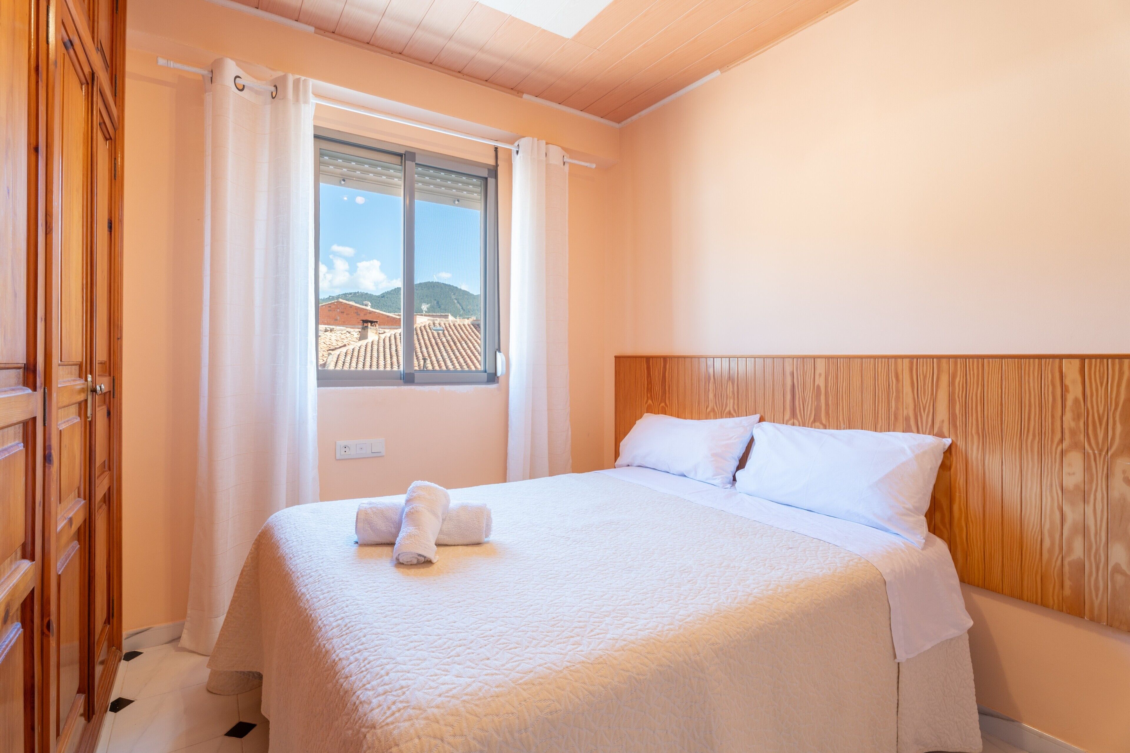 A hotel room with a bed, wooden headboard, a window with curtains, and a view of buildings outside.