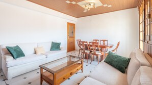 Ein Wohnzimmer mit einem weißen Sofa, grünen Kissen, einem Glastisch, Holzsesseln und einer Holzbalkendecke.