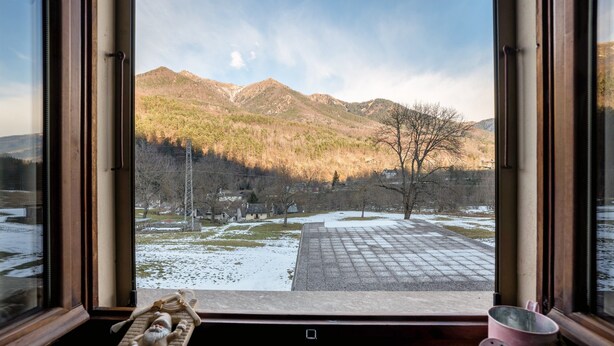 A window view of a mountainous landscape with a clear sky, a wooden table with a decorative item, and a pink mug on the windowsill. - Apartment 'Daniela' with Mountain View, Balcony and Wi-Fi (Druogno)
