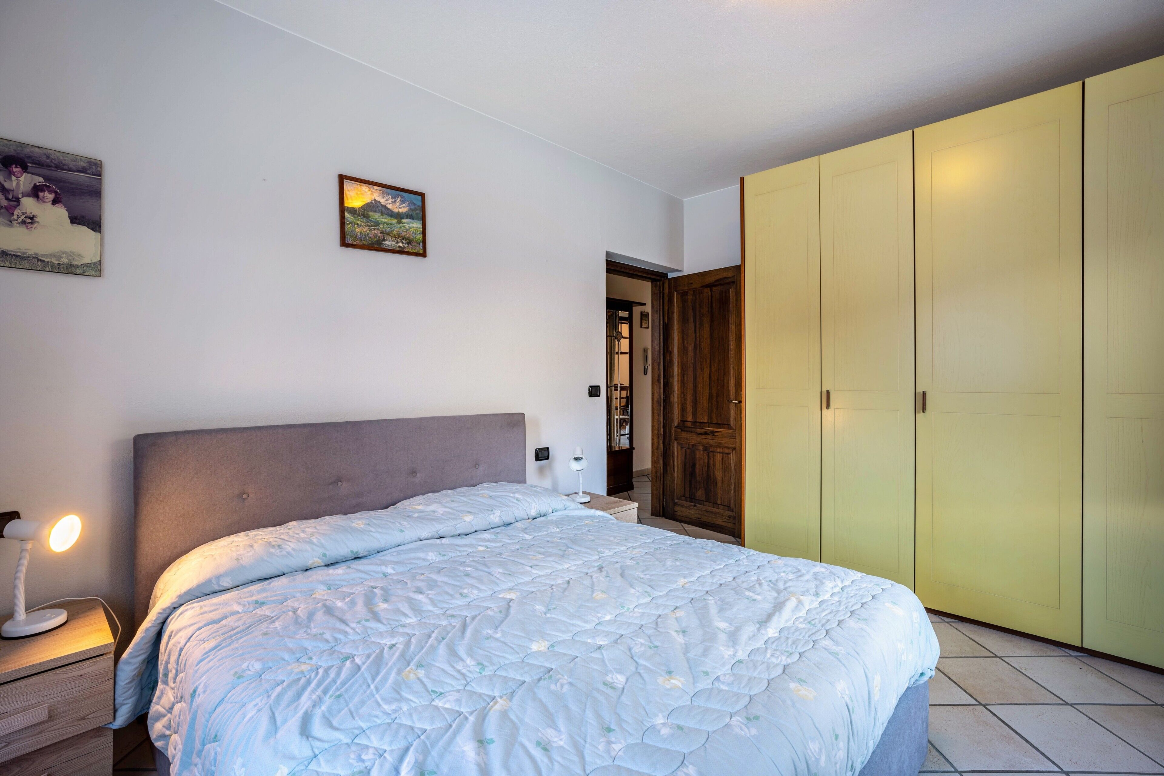 A bedroom with a bed, bedside table, lamp, wardrobe, and a painting on the wall.