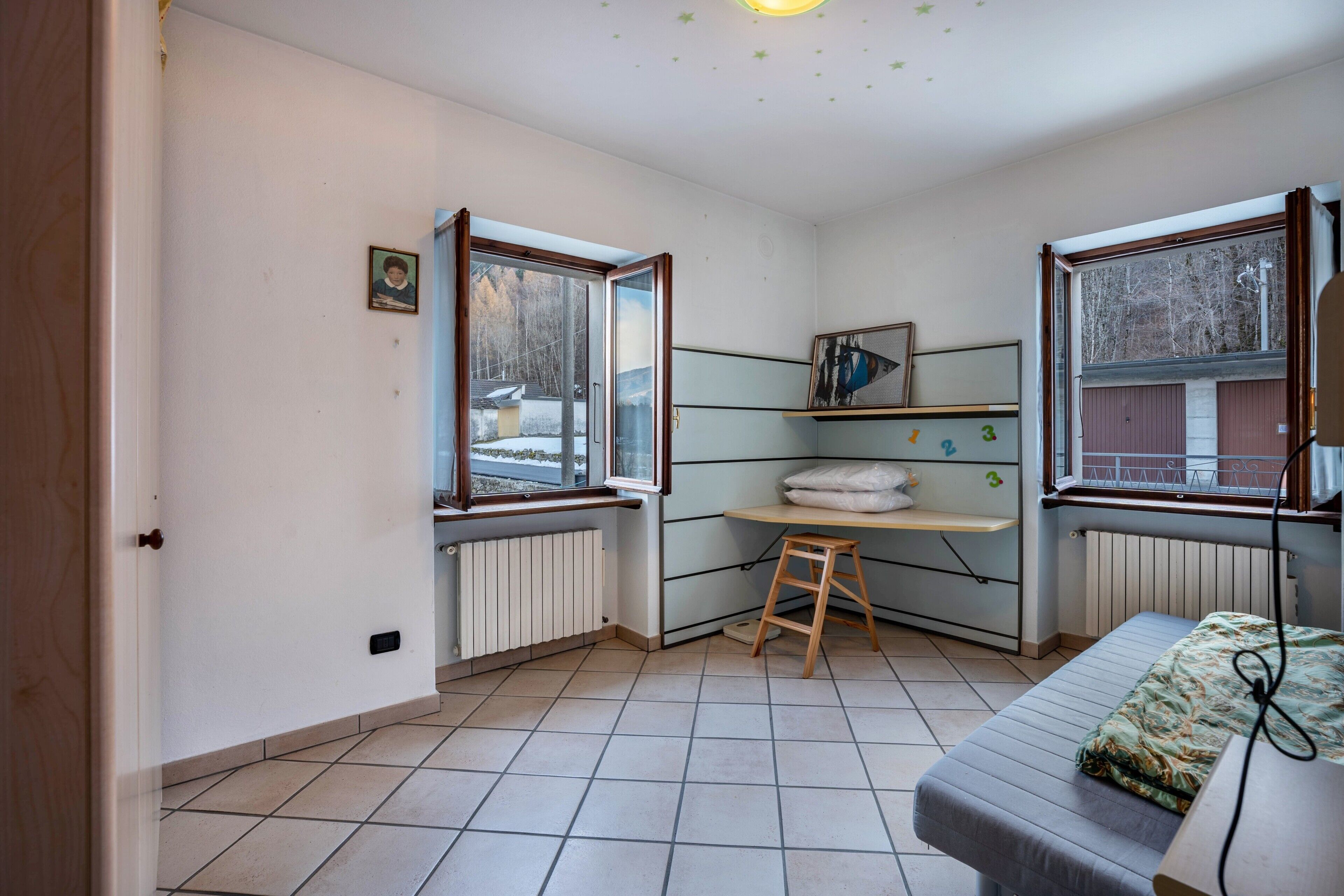 Ein Zimmer mit einem Bett, einem Schreibtisch, einer Heizung und Fenstern mit Blick auf Bäume.