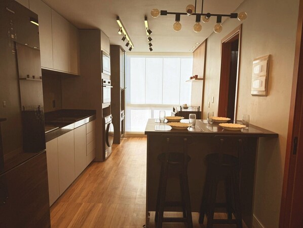 A modern kitchen with wooden cabinets, a central island with stools, and a large window providing natural light. - SaltBeach Atalaia Residence Inn - M202 (Salinópolis)