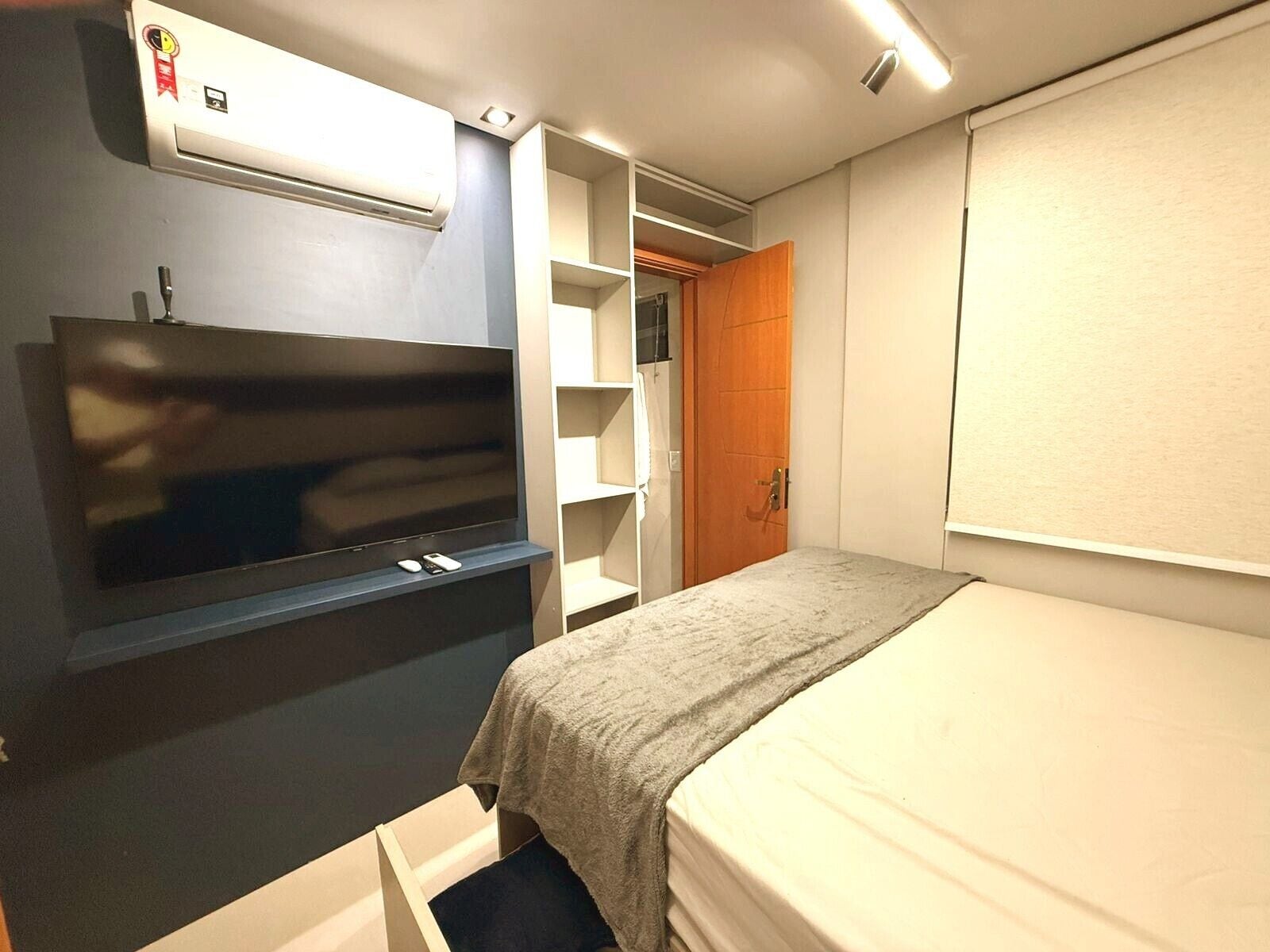 Quarto de hotel compacto com cama, televisão, guarda-roupa e ar condicionado integrado.