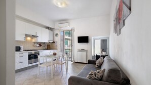 A modern kitchen with white cabinets, a dining area with chairs, a grey sofa, and a flat-screen TV mounted on the wall.