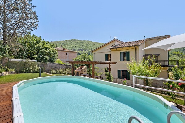 A swimming pool with a wooden deck, a pergola, and a house with a tiled roof in the background.