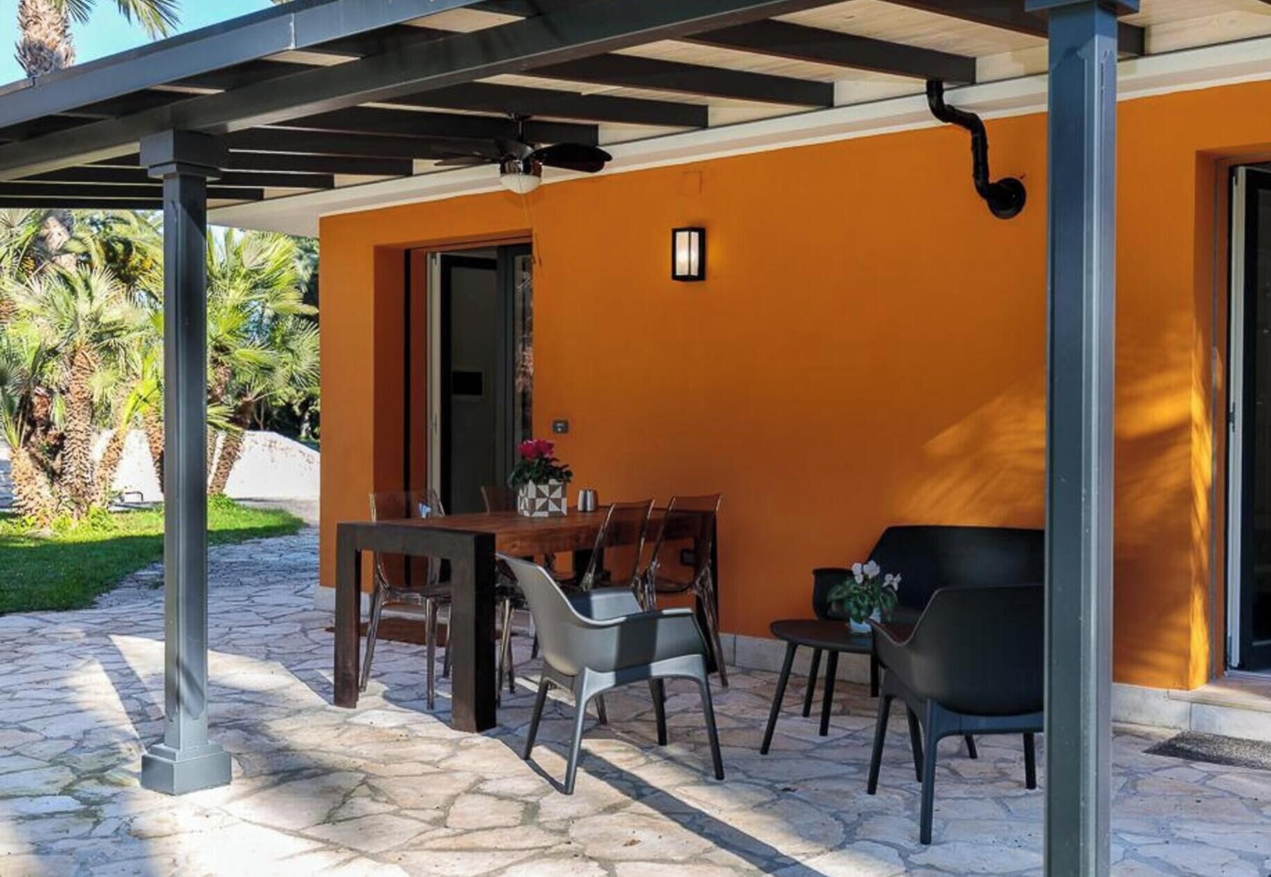 Um alpendre coberto com mesa e cadeiras, uma porta aberta e um pequeno vaso com planta.