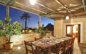 An outdoor dining area with a stone wall, wooden table, and chairs, set against a backdrop of palm trees and a clear sky.