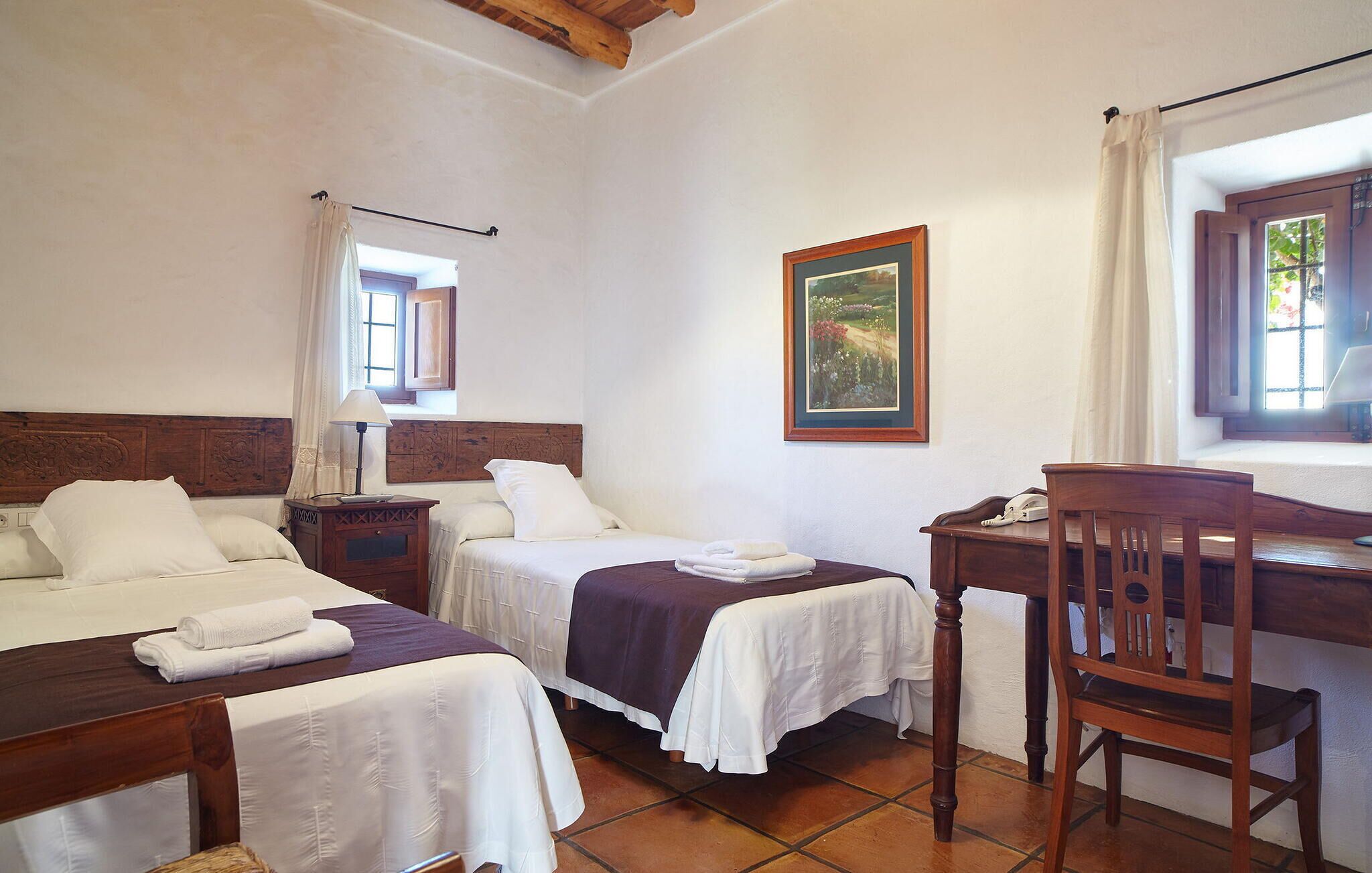 Habitació d'hotel amb dos llits, escriptori, cadira, un quadre i una finestra amb cortines.