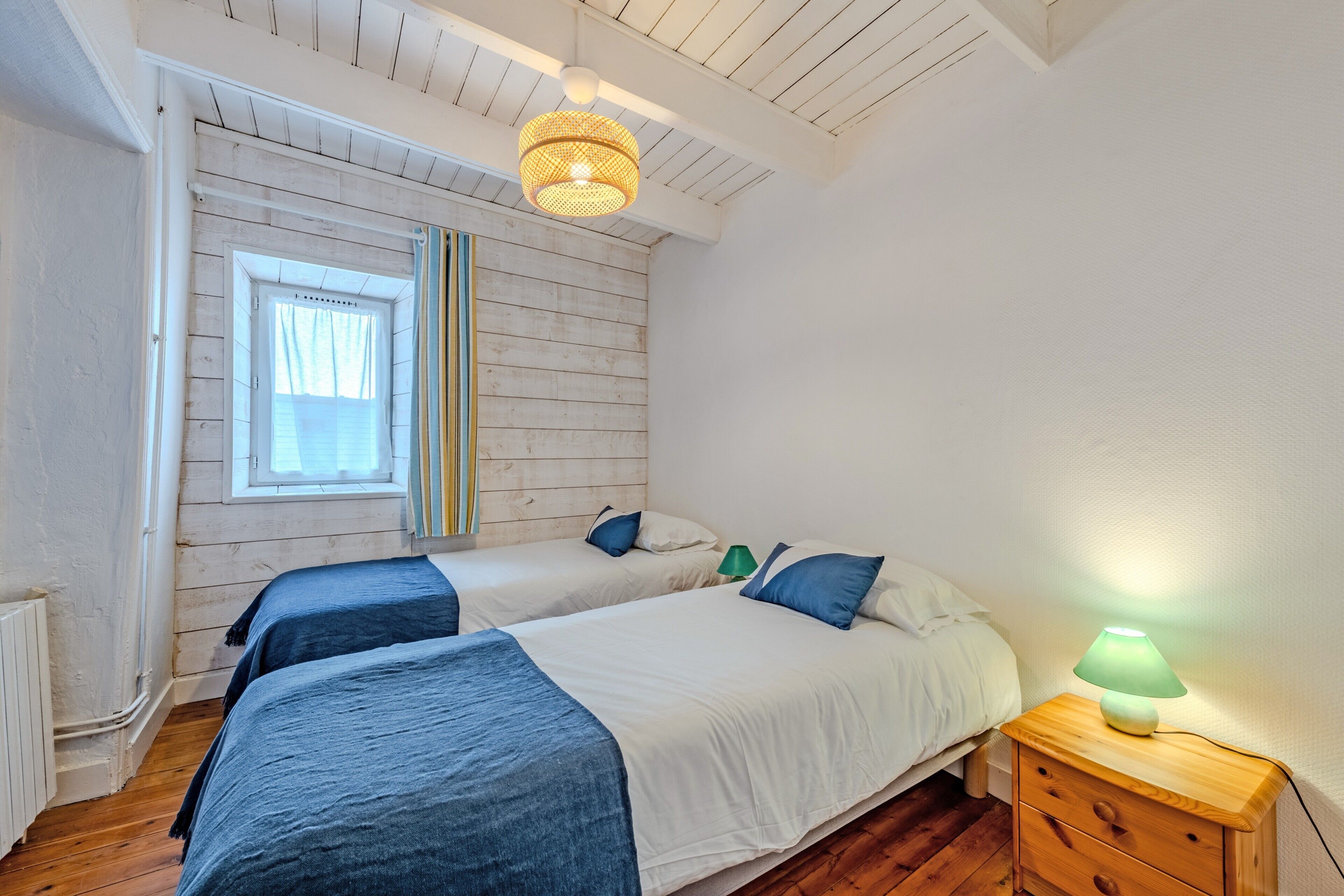 A bedroom with two beds, a window, a wooden ceiling, and a nightstand with a lamp.