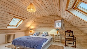 Un dormitorio con techo inclinado, paredes de madera, una cama con colcha azul, dos mesitas de noche con lámparas, una silla y un radiador.