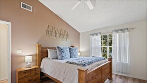 Un dormitorio con una cama de madera, una mesita de noche, una lámpara, un ventilador de techo y una ventana con cortinas.