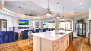 Una cocina moderna con una isla central, un comedor con sofás azules y una sala de estar con televisor y chimenea.