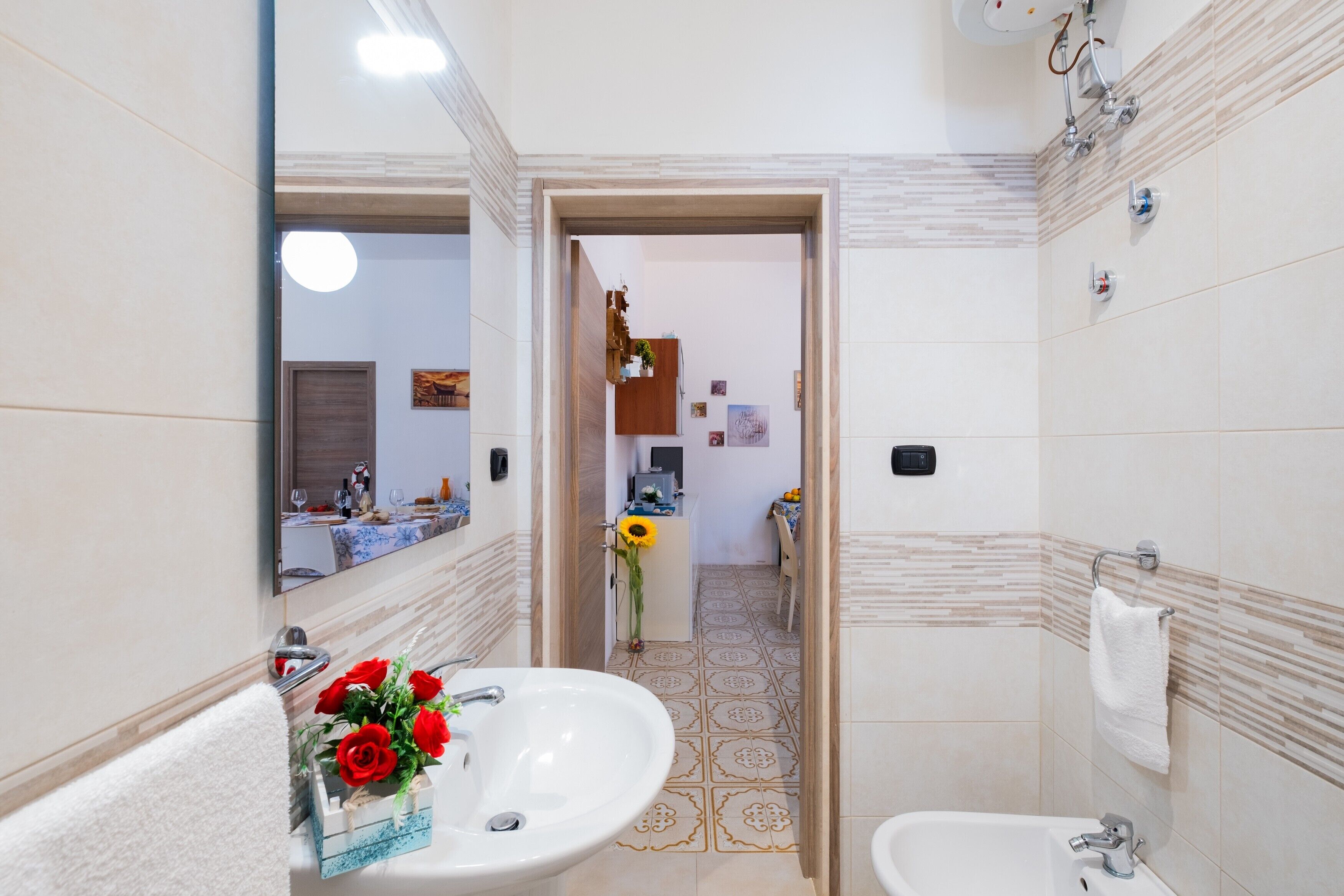 Une salle de bain avec un lavabo, un miroir et deux toilettes. Une porte donne accès à une cuisine.
