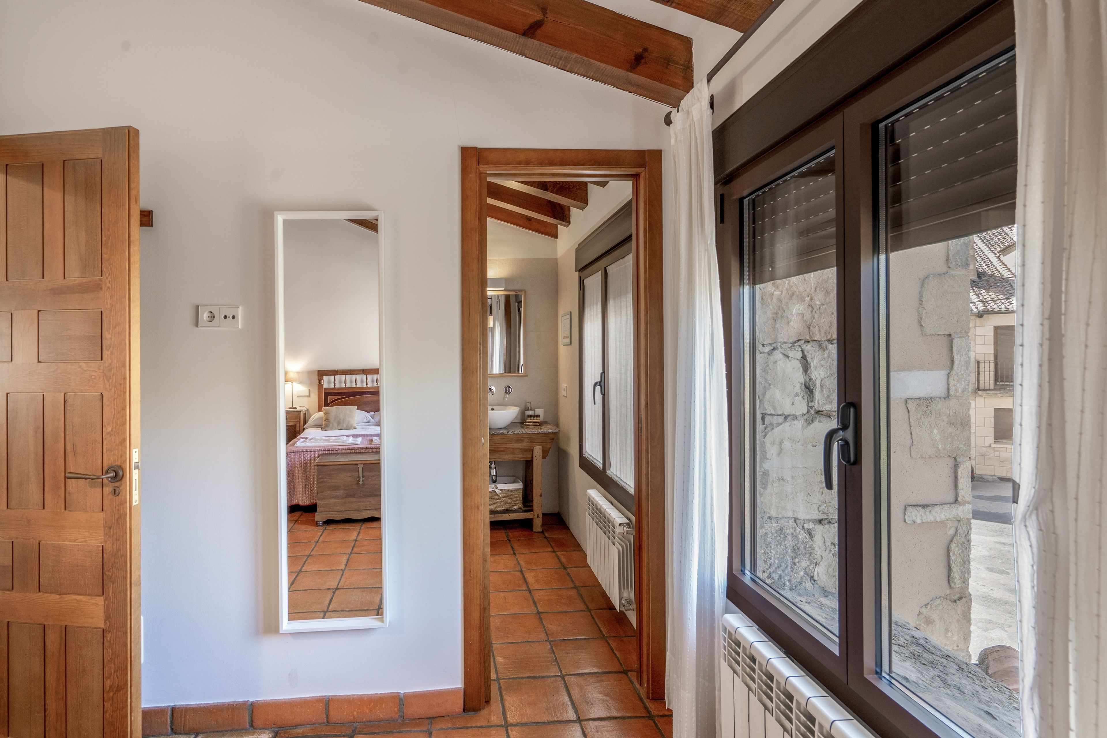 Un couloir mène à une chambre dotée d’une porte en bois, à une salle de bain avec un lavabo et un miroir, et à une fenêtre donnant sur un mur en pierre.