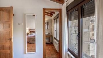 Un couloir mène à une chambre dotée d’une porte en bois, à une salle de bain avec un lavabo et un miroir, et à une fenêtre donnant sur un mur en pierre.