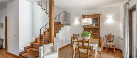 A spacious living area with a staircase, a dining table with chairs, and a kitchen area with a wooden cabinet and a hanging light fixture.