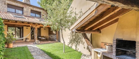 Un patio con un sendero de piedra, una pérgola de madera, una zona de barbacoa y un edificio de piedra con techo de tejas.