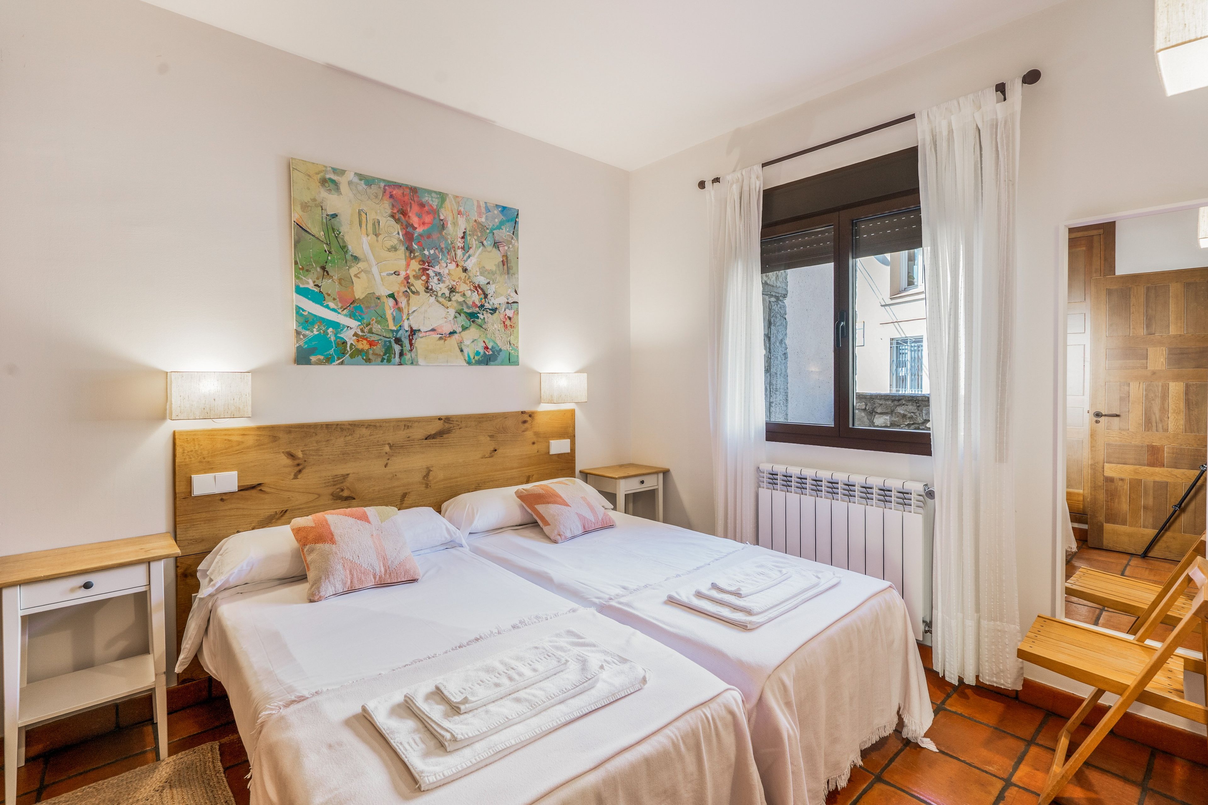 Ein Doppelbett mit weißer Bettwäsche, ein Holz-Kopfteil, zwei Nachttische, ein farbenfrohes abstraktes Gemälde, ein Fenster mit Vorhängen und ein Heizkörper.