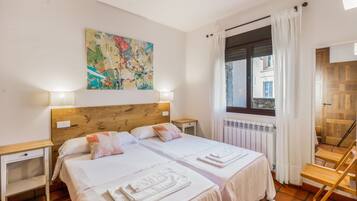 Ein Doppelbett mit weißer Bettwäsche, ein Holz-Kopfteil, zwei Nachttische, ein farbenfrohes abstraktes Gemälde, ein Fenster mit Vorhängen und ein Heizkörper.