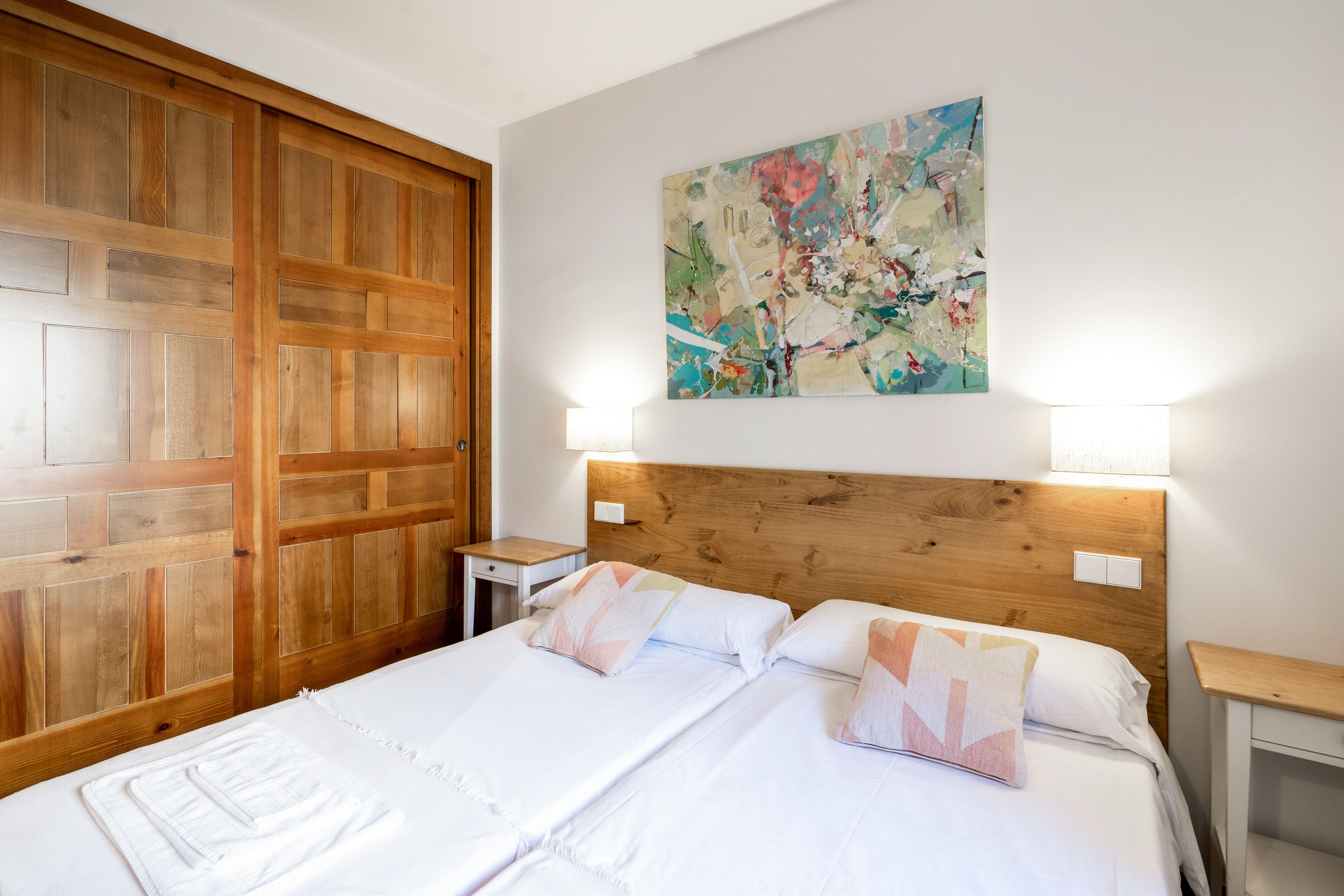 Cama doble con sábanas blancas y dos almohadas, cabecera de madera, mesita de noche con lámpara y armario de madera.