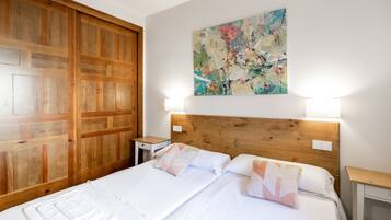 Cama doble con sábanas blancas y dos almohadas, cabecera de madera, mesita de noche con lámpara y armario de madera.