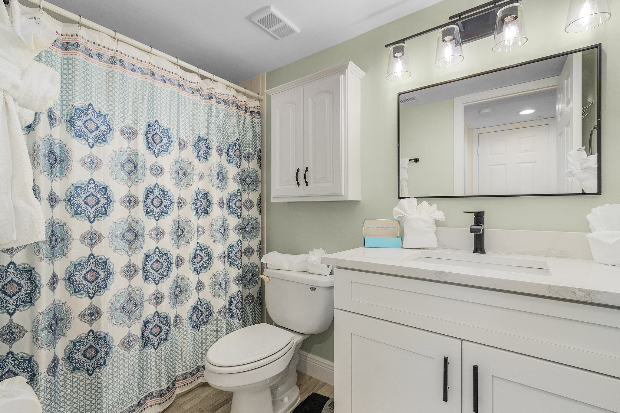 Un baño con cortina de ducha estampada, inodoro, lavabo con espejo y armarios.