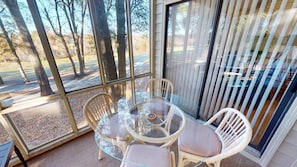 A glass-enclosed patio with wicker chairs and a glass table, overlooking a wooded area. - River Creek Golf Condo | Your Ultimate NC Escape! | RC 303 (Sunset Beach)