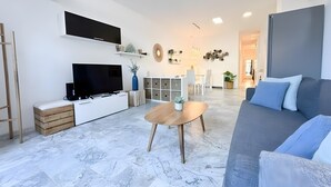 A modern living room with a flat-screen TV, a wooden coffee table, a gray sofa, and a dining area with white chairs. - Apartment 'Ola De Mar' with Shared Pool, Wi-Fi and Air Conditioning (Estepona)