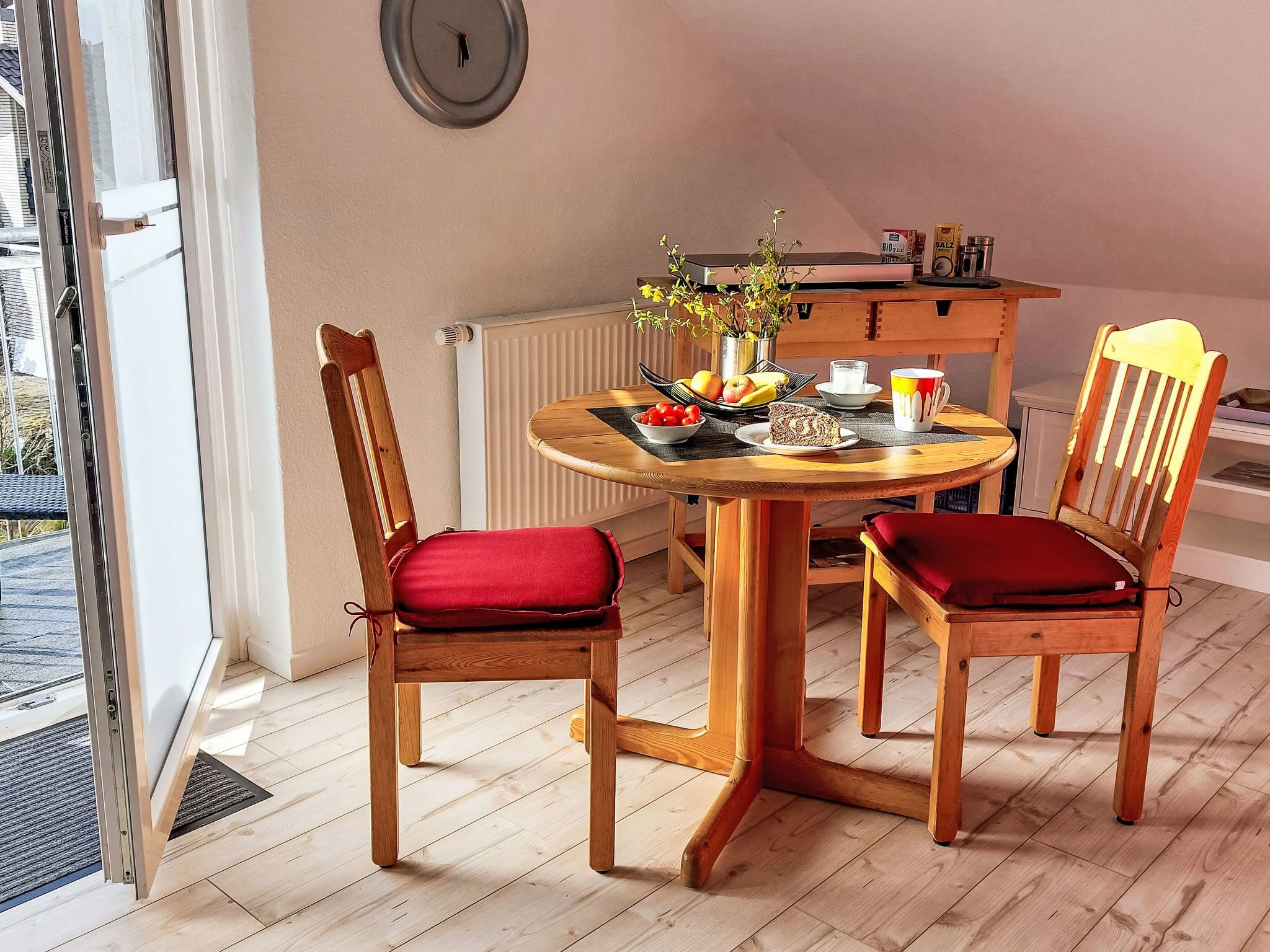 Ein Essbereich mit einem runden Holztisch, zwei Holzsesseln mit roten Kissen, einer kleinen Topfpflanze und einer Uhr an der Wand.