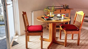 Un coin repas avec une table ronde en bois, deux chaises en bois avec des coussins rouges, une petite plante en pot et une horloge au mur.