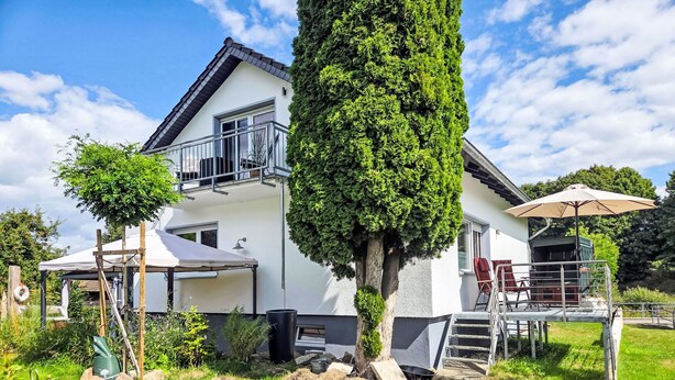 Une maison à deux étages avec un balcon, une terrasse couverte et un grand arbre dans le jardin.