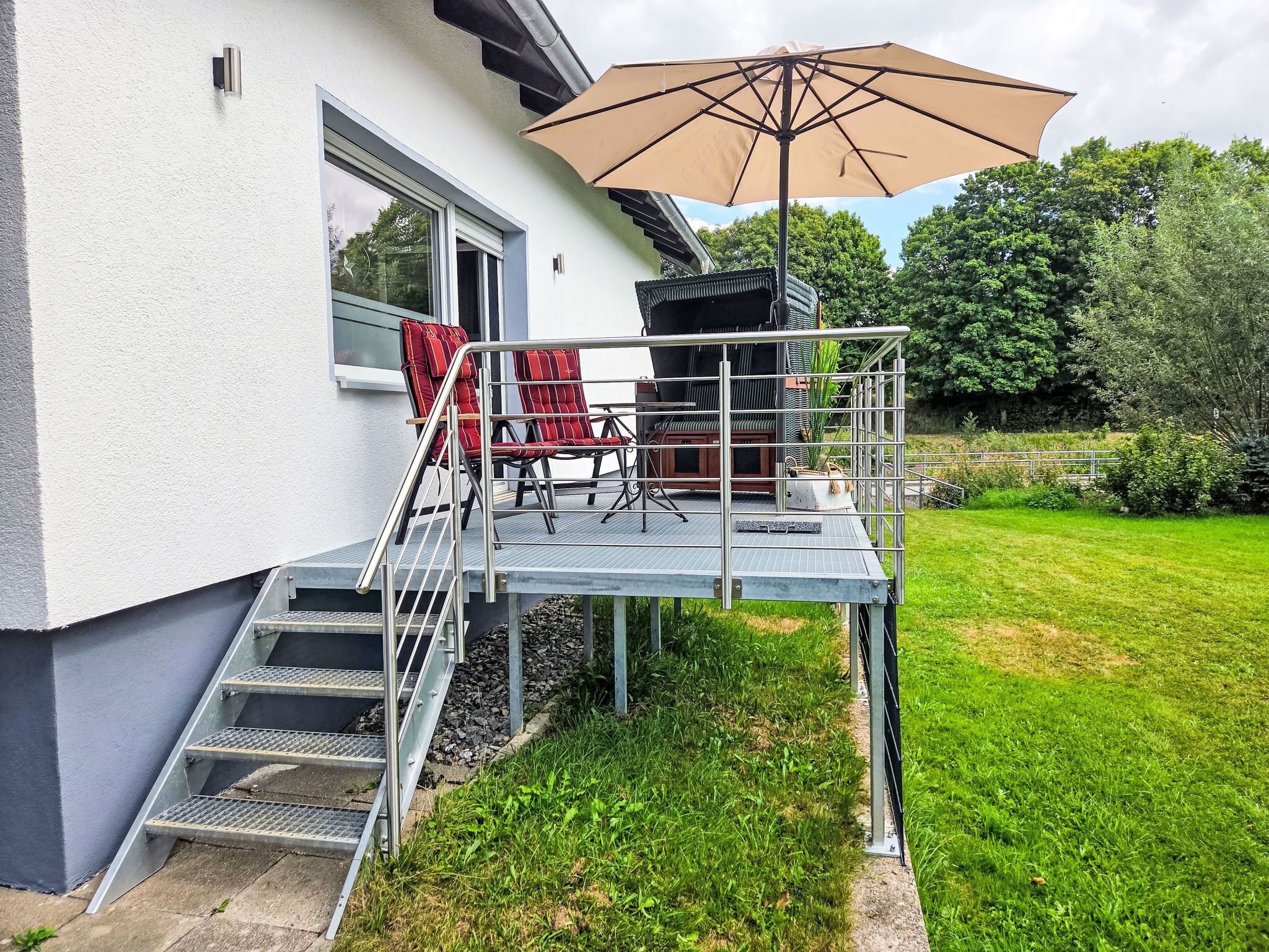 A modern house with a covered outdoor seating area, a staircase leading to the entrance, and a well-maintained lawn.