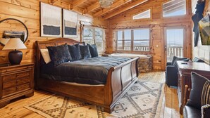 Une chambre aménagée dans un chalet en rondins, avec un grand lit, un bureau en bois, une chaise, une table de chevet, une lampe, un lustre et une cheminée en pierre.