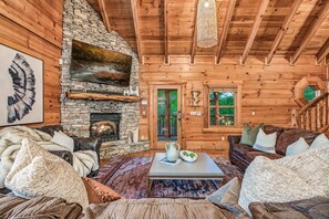 L’intérieur d’une cabane en rondins, avec une cheminée en pierre, des meubles en cuir et une vue sur l’extérieur.