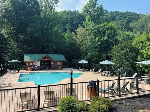 A swimming pool with lounge chairs and umbrellas, surrounded by a fence, and a small building with a green roof.