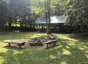 A rustic outdoor area with a metal-roofed structure, two wooden benches, and a fire pit surrounded by trees.