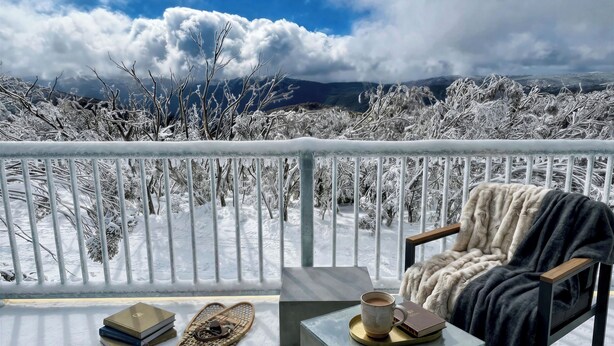 Un balcón con vistas a árboles nevados y montañas, una silla con una manta, y una mesa con libros y una taza.