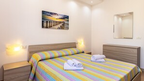 Une chambre d’hôtel avec un lit, une table de chevet, une lampe de chevet, un miroir fixé au mur et un tableau représentant un coucher de soleil.