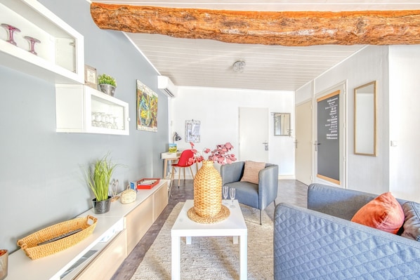 Ein modernes Wohnzimmer mit einem weißen Couchtisch, einem blauen Sofa und einem grauen Schlafsofa. Man sieht einen Holzbalken an der Decke, ein an der Wand befestigtes Regal mit Dekorationen sowie eine Tür mit einem Schild.