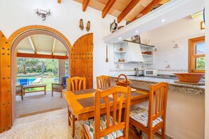 A wooden dining table with chairs, a kitchen with white cabinets and a microwave, and an outdoor area with a pool and chairs.