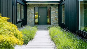 A modern building with a stone facade, a dark green door, and a pathway flanked by green and purple plants.