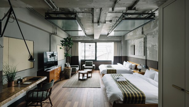 A modern hotel room with a wooden floor, a bed with a plaid blanket, a desk with a computer, a TV, and a large window with curtains.