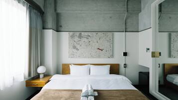 A modern hotel room with a large bed, a wooden nightstand, a wall-mounted lamp, and a large window with curtains.