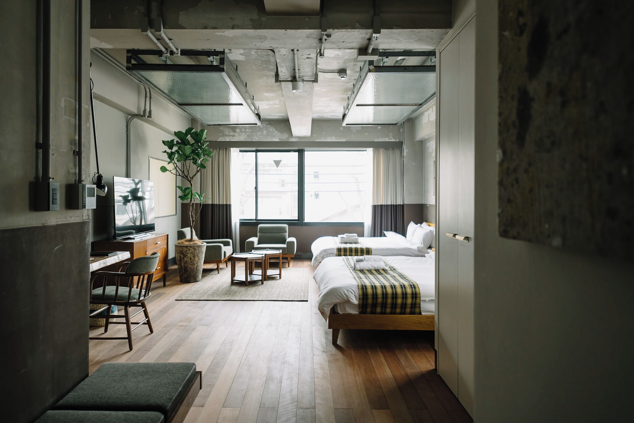 Une chambre d’hôtel moderne avec un plancher en bois, un lit, un bureau avec une chaise, une télévision et une grande fenêtre avec des rideaux.
