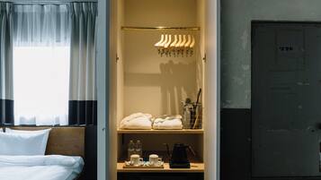 Ein Hotelzimmer mit Bett, Kleiderschrank mit Kleidung, Ablagefläche mit Handtüchern und Pflegeprodukten sowie Fenster mit Vorhängen.