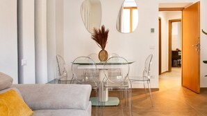 Een moderne eetruimte met een glazen tafel en transparante stoelen, een vaas met gedroogde planten, en een houten deur op de achtergrond.