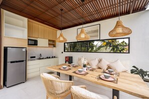 A modern kitchen with wooden cabinets, a stainless steel refrigerator, and a dining area with a wooden table set for two. - Jing Jang Villa Ungasan (Kutuh)