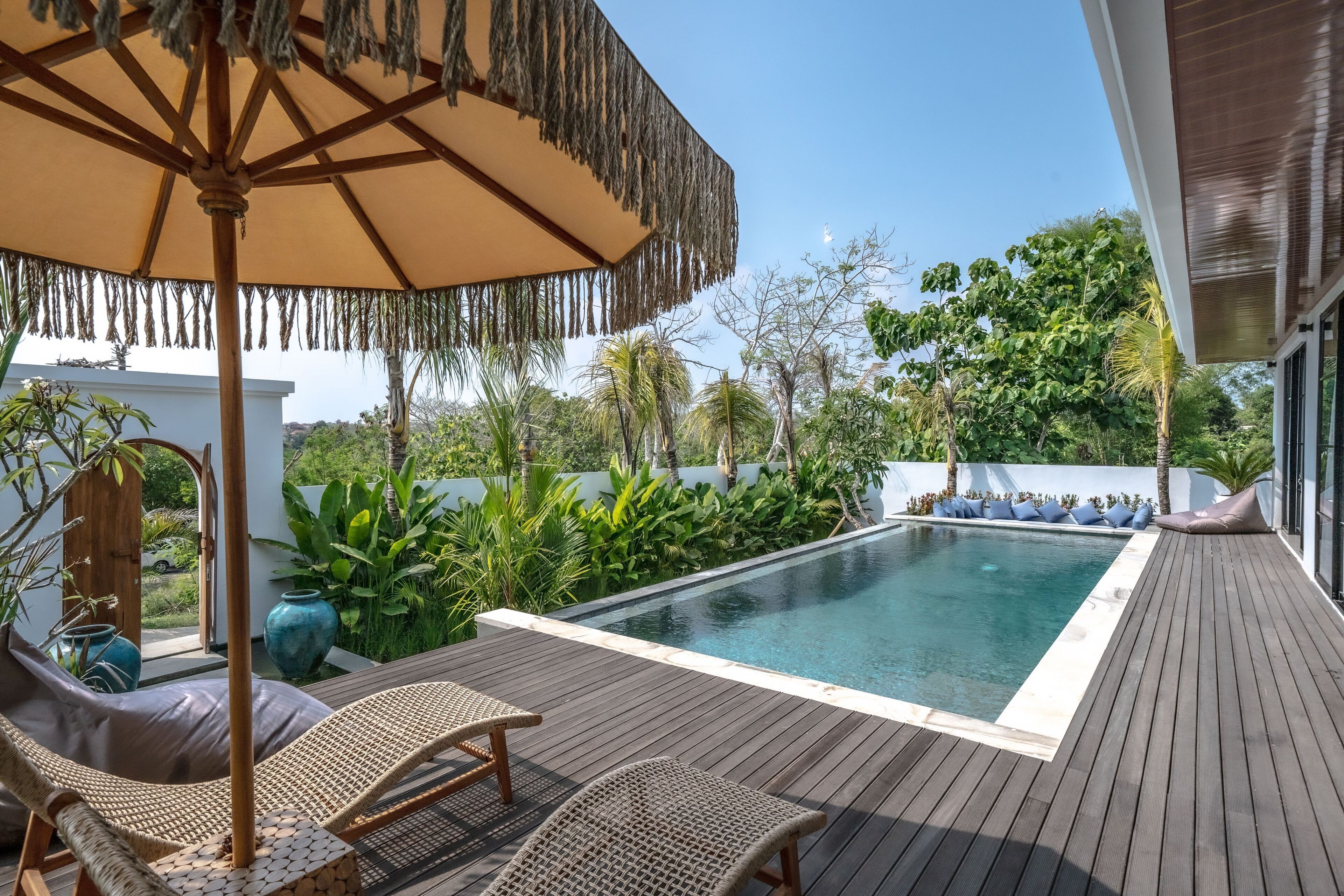Um deck de madeira com piscina, cadeiras de sol e um guarda-sol de palha.