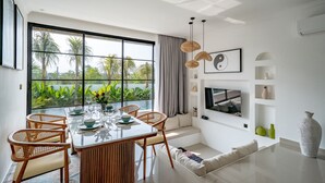 Ein modernes Wohnzimmer mit einem großen Fenster, einem Essbereich mit Korbstühlen, einer Couch und einem an der Wand montierten Fernseher.